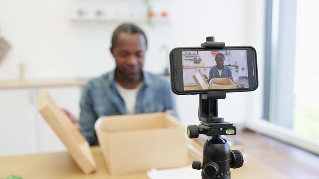 An enthusiastic creator records a home unboxing video on a smartphone tripod, with an open cardboard package and bright kitchen behind him