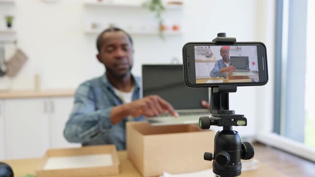 A creator records a hands-on presentation in a bright home workspace, using a smartphone on a tripod while cardboard boxes and a laptop sit nearby