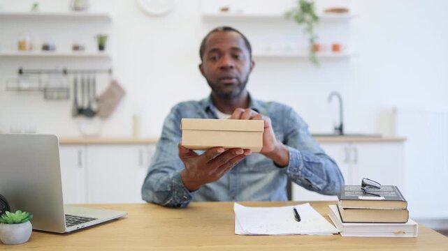 A smiling man raises his fist while holding a cardboard parcel at a wooden table, with a laptop, loose papers, and stacked books in a bright home interior