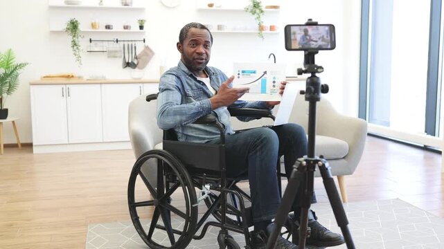 A wheelchair user shares a printed data chart during a home video presentation, with a laptop and smartphone recording setup in a bright kitchen