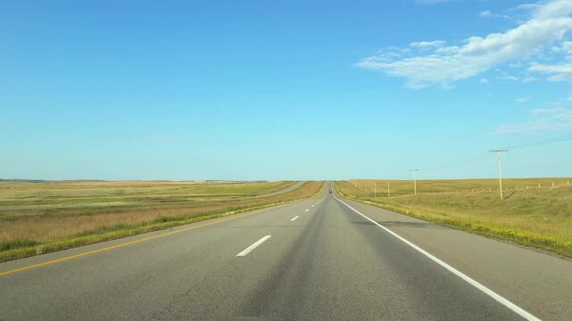 Point of view driving on empty two-lane highway through dry grass prairie farmland with power lines in Saskatchewan. 