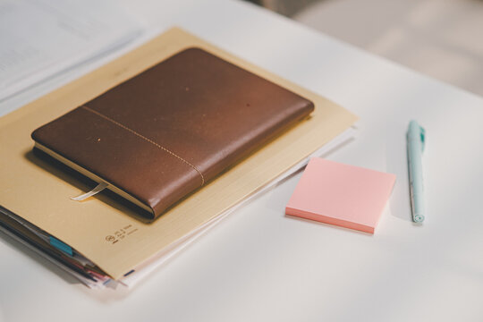 Brown leather notebook lying on folder document with pink sticky notes and pen on white office desk.