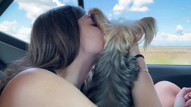 Young woman enjoying a beautiful car ride and affectionately kissing her cute Yorkshire Terrier dog while looking out the window, sharing moments of love and pleasure