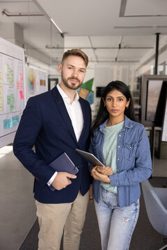 Confident diverse office employees posing for portrait at modern workspace holding digital and paper work tools looking at camera as competent and reliable professional corporate staff representatives