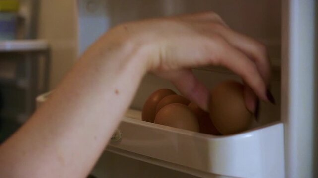 Close-up captures hand reaching for brown chicken eggs stored in refrigerator door compartment; soft lighting emphasizes texture and natural hue.

