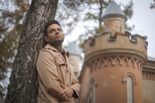 Thoughtful man in trench coat leaning on tree near ancient castle tower with copy space
