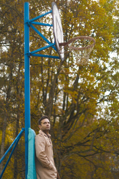 Thoughtful man in trench coat standing near basketball hoop in autumn park with copy space