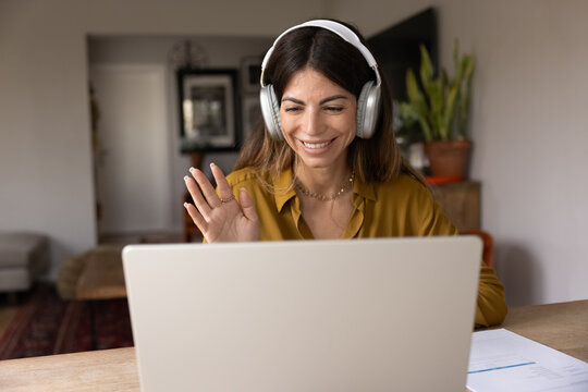 Smiling Hispanic woman in headphones waving at laptop during video call, working remotely from home office, starting virtual meeting by business. Digital communication, modern technology, app use