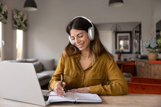 Smiling adult woman in headphones studying at home using laptop, writing notes, making exercise, developing professional skills and knowledge, listening audio course, attend online training or webinar