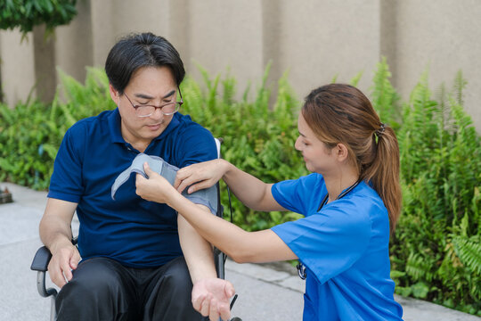 Asian female nurse measuring blood pressure of senior male patient in a wheelchair outdoor.