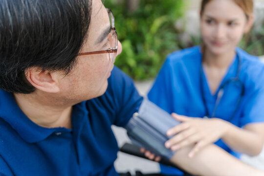 Asian female nurse measuring blood pressure of senior male patient in a wheelchair outdoor.