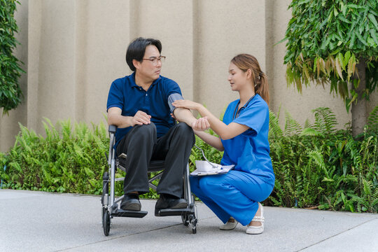 Asian female nurse measuring blood pressure of senior male patient in a wheelchair outdoor.