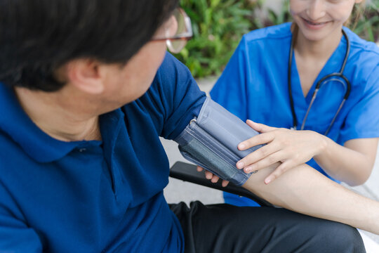 Asian female nurse measuring blood pressure of senior male patient in a wheelchair outdoor.