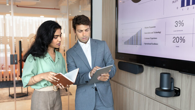 Colleagues stand next to screen displaying charts, analyzing financial data, reviewing statistics on digital tablet during collaborative meeting in office. Business analytics, strategy development