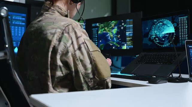 Close-up of a military officer in camouflage uniform writing on a clipboard in a high-tech command center. Glowing monitors with radar and maps are