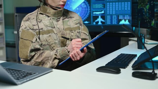 Close-up of a military officer in camouflage uniform writing on a clipboard in a high-tech command center. Glowing monitors with radar and maps are