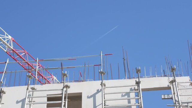 Steel rebar on a concrete wall and a red and white tower crane jib with hook against a blue sky with a contrail, French Riviera, France.