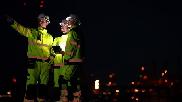 Three oil and gas engineers in white hard hats study a glowing tablet at night, one pointing up toward the plant while the bearded engineer smiles, refinery lights far behind
