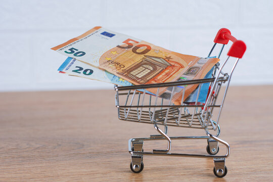 Miniature shopping cart with euro banknotes inside on wooden table. Inflation, rising cost of living, retail expense and consumer budget concept.