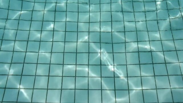 Top view of a swimming pool with light blue square tiles and crystal clear water with sun light reflections and ripples on the surface creating caustics patterns.