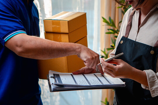A courier points to a delivery form as a worker holds a pen and clipboard. Stacked parcels behind them illustrate shipment checks, delivery confirmation, shipping paperwork, and order handover.