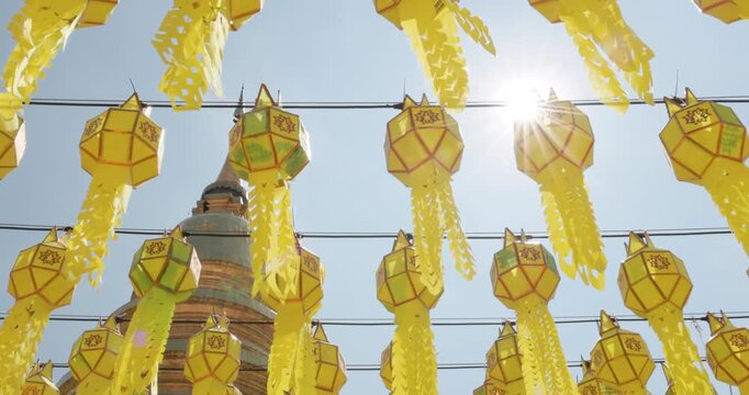 Bright yellow hanging lanterns and intricate streamers glow in sunlight before golden stupa at Wat Phra That Hariphunchai, Lamphun, Thailand, during 2026 lantern festival, low angle view with sunburst