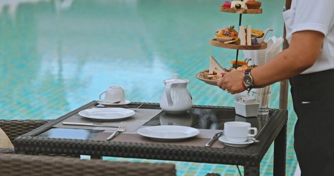 Waiter serving afternoon tea set on a tiered stand at a poolside table. Luxury resort hospitality service with snacks, sandwiches, and tea for guests relaxing by the swimming pool, slow motion.