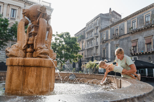 Father helps his daughter reach the fountain water in a city square