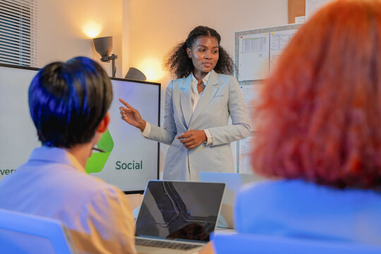 Confident businesswoman with curly hair presenting social responsibility pillar of ESG sustainability strategy on screen to colleagues during evening office meeting