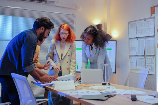 Multiethnic business team standing around meeting table and reviewing ESG sustainability documents and charts together while planning green strategy in office at night