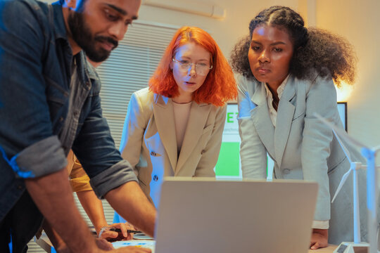 Serious multiethnic colleagues looking at laptop screen and analyzing ESG data with concern during late night sustainability meeting with wind turbine model on desk