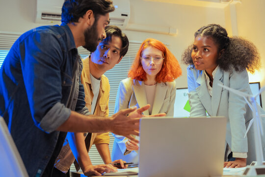 Serious multiethnic colleagues looking at laptop screen and analyzing ESG data with concern during late night sustainability meeting with wind turbine model on desk