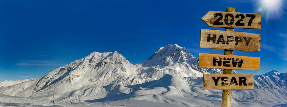 happy new year 2027 written on a wooden directional sign with a panoramic view of the snowy peaks of a ski area in the Alps background
