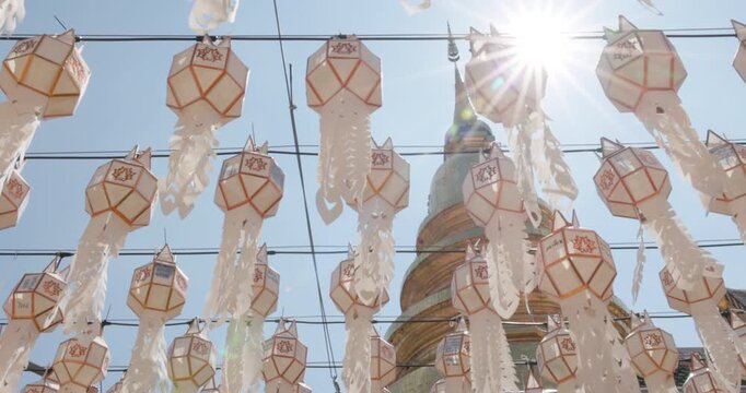 Rows of white paper lanterns with long cutout streamers hang before golden stupa at Wat Phra That Hariphunchai, Lamphun, Thailand, during 2026 lantern festival, sunburst shines through decorations