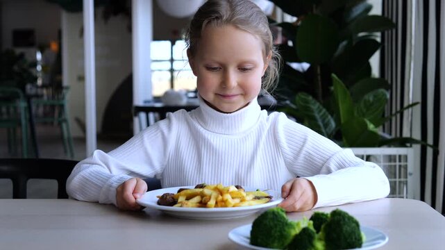 Young girl refuses broccoli at lunch, frowning as she looks at green vegetables beside plate of fries. Hesitant child pulls her meal closer, showing food refusal in bright casual dining room.