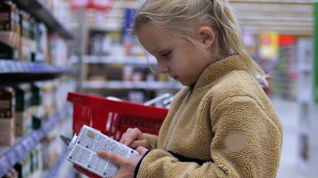 Child studies package label in grocery aisle, holding item beside red shopping basket. Attentive young shopper compares product details during everyday family shopping outing, reading label