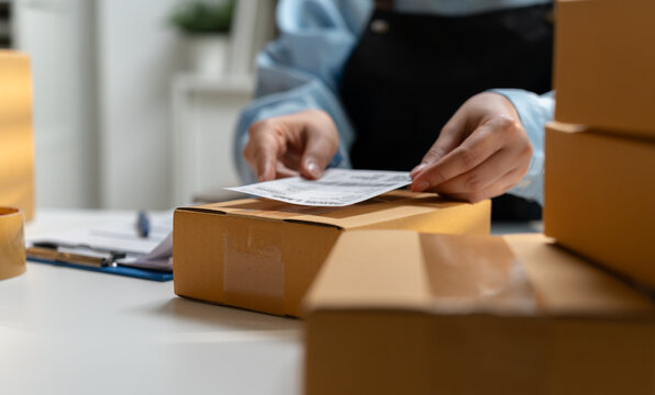 Close-up of a small business worker placing a shipping label on a cardboard package at a packing desk, illustrating ecommerce order fulfillment, parcel preparation, logistics, and delivery operations.
