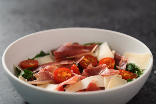 Salad with prosciutto, tomato, arugula leaves and parmesan cheese in white bowl on concrete surface