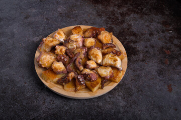 Traditional Galician-style octopus served with boiled potatoes, olive oil and paprika on a round wooden plate against a dark textured background. Spanish cuisine © patxi