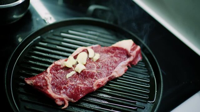 Raw beef steak with garlic cloves on a round cast iron grill pan as a sprig of rosemary is added, French Riviera, France.