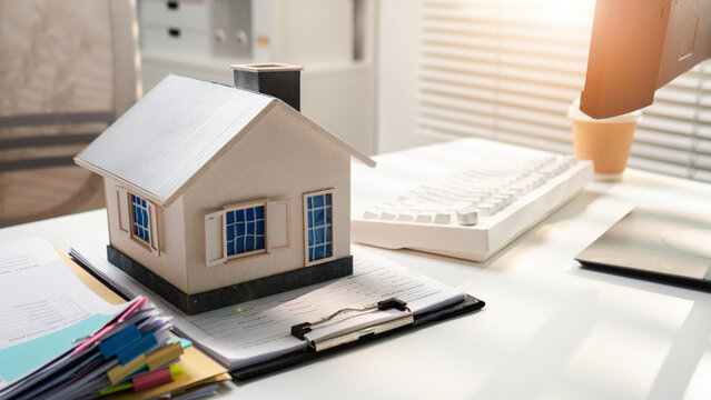 A miniature house sits on property paperwork beside a desktop computer. The sunny office scene illustrates home buying, mortgage applications, real estate contracts, and investment planning.