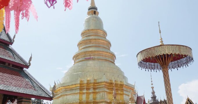 Pink and orange hanging lantern streamers frame golden stupa and ornate parasol at Wat Phra That Hariphunchai, Lamphun, Thailand, during 2026 lantern festival, low angle view beneath clear sky