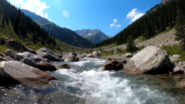 A clear stream cascades through a valley surrounded by mountains, rocks, and trees, creating a scenic landscape under a blue sky with some clouds