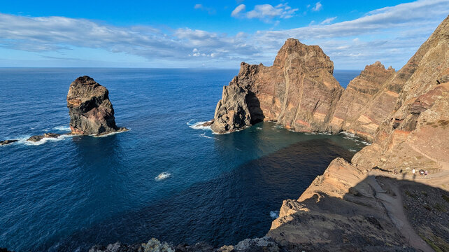 Layered red and ochre volcanic cliffs and a sea stack rising from deep blue Atlantic water, with hikers on the coastal trail, at Ponta do Castelo, Ponta de S&atilde;o Louren&ccedil;o, Madeira, Portugal.