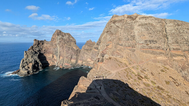 Layered red and ochre volcanic cliffs and a sea stack rising from deep blue Atlantic water, with hikers on the coastal trail, at Ponta do Castelo, Ponta de S&atilde;o Louren&ccedil;o, Madeira, Portugal.