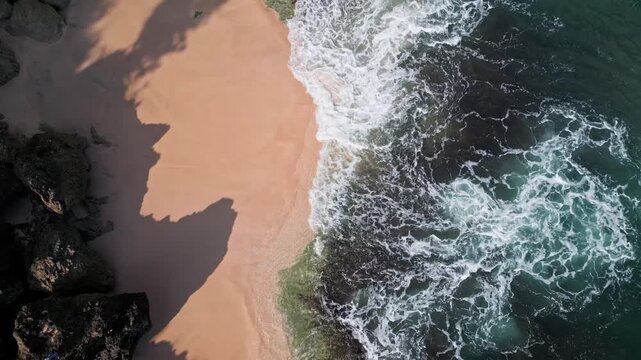 Aerial bali coastline rocky beach waves washing over golden sand and dark volcanic rocks, turquoise water swirling into white foam, dramatic shadows from cliffs, soothing rhythmic, cinematic video