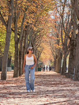 An Asian woman strolls along a leaf-covered path beneath golden autumn trees in Paris, France, on a sunny day, with other visitors in the distance. Jardins des Champs Elysees