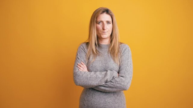 Stern middle aged woman crossing her arms and looking at the camera with a disapproving expression. Serious lady judging someone over a yellow background