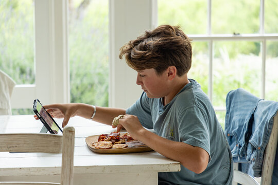 13 year old boy eating pancakes and looking at tablet computer in light filled room early morning