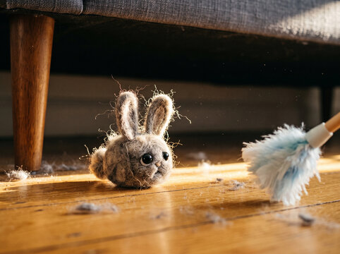 Cute needle felted dust bunny with big black eyes hiding under a sofa in a sunbeam as a blue feather duster approaches, spring cleaning concept